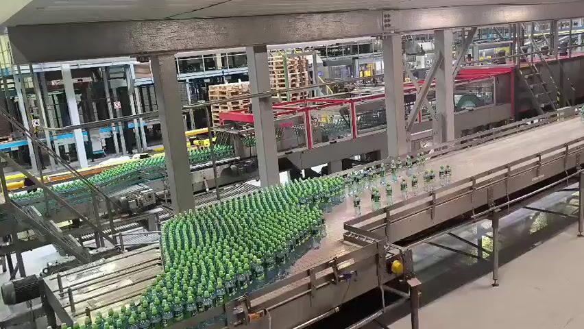 The video shows a wide shot of a bottling plant with a conveyor belt system. Thousands of green bottles are moving along the conveyors. Some bottles are in a large formation, while others are in a single line. A person wearing a yellow safety vest and a white hard hat is visible on the right side of the frame, adjusting their hard hat. The environment appears to be a large industrial facility with metal structures and machinery. The lighting is bright, suggesting an operational setting. The video captures the automated process of bottle transportation within the plant.