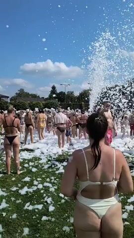 The video shows a large outdoor foam party on a sunny day with a blue sky and white clouds. Many people, mostly adults and some children, are dressed in swimwear and dancing in a grassy area covered in a thick layer of white foam. In the foreground, a woman wearing a light-colored one-piece swimsuit faces away from the camera. In the midground and background, more people are visible, some interacting with the foam and each other. A powerful foam machine is in the background, spraying a large amount of foam into the air. The overall scene depicts a lively and celebratory atmosphere.