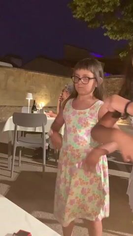 The video shows a young girl with dark hair and glasses, wearing a light-colored floral dress, dancing in what appears to be an outdoor dining area at night. She is in the foreground, with her arms moving. Behind her, another person, mostly obscured, is also visible, their arms in motion. In the background, tables with white tablecloths, chairs, and some lighting are present, along with a stone wall and trees. The overall lighting suggests it is evening or night.