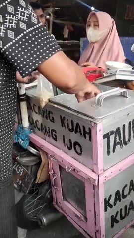 A person with a black and white patterned shirt is holding a clear plastic bag with small items inside. They are reaching into a metal container on a pink and silver cart. The cart has writing on it that appears to say "KACANG KUAH IJO" and "TAUW" and "KACANG KUAH". Another person wearing a pink headscarf and a mask is standing behind the cart. A man wearing a grey shirt is standing behind the woman. A motorcycle is parked next to the cart. The environment appears to be outdoors, possibly a market.