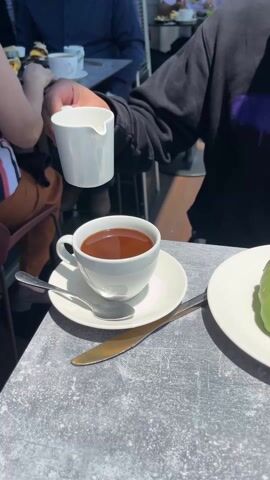 A person pours cream into a cup of coffee sitting on a grey concrete table. Next to the cup, on a white saucer, lies a silver spoon and gold knife.  To the right, there’s a white plate with a croissant dipped halfway in green frosting with pistachio nuts on top. A person in a black sweatshirt is pouring the cream. Other people sit in the background at the table.