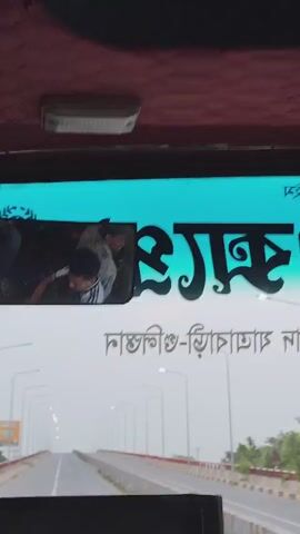 The video appears to be filmed from inside a bus, looking out through the windshield. The perspective is a first-person view of the road ahead, suggesting the bus is in motion.

**Subjects:**
*   **People:** In a reflection on the upper part of the windshield, two young men are visible. One is wearing a white shirt with black stripes and appears to be interacting with the other. Their expressions are difficult to discern clearly due to the reflection and resolution.
*   **Objects:** The primary objects are the interior of the bus (dashboard, windshield, interior light), the road, a long bridge with streetlights lining both sides, a billboard on the left side of the road, and barriers along the sides of the bridge.
*   **Scene/Setting:** The scene is a bridge, likely a highway or major road, extending into the distance. The sky is overcast and hazy, suggesting a cloudy or foggy day. The environment appears to be rural or semi-rural, given the open road and what looks like vegetation on the sides. The presence of a long bridge indicates a crossing over water or a valley.

**Activity or Event:**
The bus is traveling across a bridge. The video captures the forward motion and the view of the road ahead. The interaction between the two individuals in the reflection adds a human element, possibly a brief moment of camaraderie or conversation within the bus.

**Notable Details:**
*   **Time of Day/Weather:** The lighting suggests it is daytime, but the hazy conditions make it difficult to determine the exact time. The weather appears to be overcast or foggy.
*   **Location Cues:** The text on the windshield, which reads "Kashiani-Gopalganj" (though partially obscured), clearly indicates the location context as Kashiani, Bangladesh. The style of the bus interior and the visible road infrastructure are also characteristic of the region.
*   **Emotions:** No strong emotions are evident from the visible subjects. The overall mood conveyed is one of travel and observation.

**Visible Text:**
*   On the upper part of the windshield, in large, decorative black lettering, is the word "হায়দার" (Haydar) or a similar Bengali name.
*   Below that, in smaller Bengali script, is "কাশিয়ানি-গোপালগঞ্জ" (Kashiani-Gopalganj).
*   On a billboard to the left, there is an image of a person and some Bengali text, but it is too small and blurry to read.