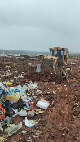 The video shows a yellow bulldozer stationary amidst a large garbage dump under an overcast sky. The ground is muddy, with clear tire tracks leading up to the bulldozer. The surrounding area is covered in a mix of plastic bags, boxes, and other types of waste. A person is visible in the driver's seat of the bulldozer.