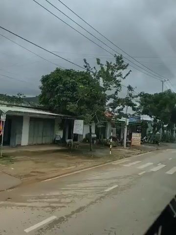 The video shows a roadside view of a commercial area in what appears to be Southeast Asia, possibly Vietnam, under a cloudy sky. The camera is positioned from a moving vehicle, likely a bus or car, offering a continuous shot of the street and buildings.

In the foreground, a pedestrian crossing is visible, marked with white stripes. The road is wet, suggesting recent rain or damp conditions. Overhead power lines crisscross the frame, supported by utility poles.

Along the roadside, several single-story buildings house various businesses. One building displays signs with Vietnamese text, and some appear to be shops or eateries. Motorbikes are parked outside some establishments, a common mode of transportation in the region. A person on a motorbike carrying goods is seen driving on the road.

Trees line parts of the street, with lush green foliage providing some contrast to the muted colors of the buildings and overcast sky. In the distance, hills or mountains can be seen. The overall atmosphere is one of a typical day in a developing urban or semi-urban area, with everyday activities taking place along the road.