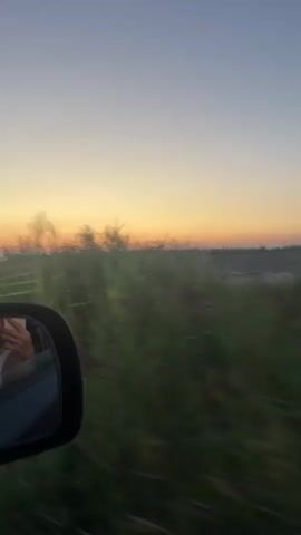 The video shows a view from inside a moving vehicle, looking out the side window and into the side mirror. The side mirror reflects a person holding a phone, presumably taking the video. Outside the vehicle, tall green grass is blurred due to motion, suggesting the vehicle is traveling at a good speed. The horizon features a sunset with warm orange and yellow hues against a pale blue sky. In the distance, silhouetted structures and a truck can be seen against the sunset. The overall impression is one of a scenic drive at dusk.