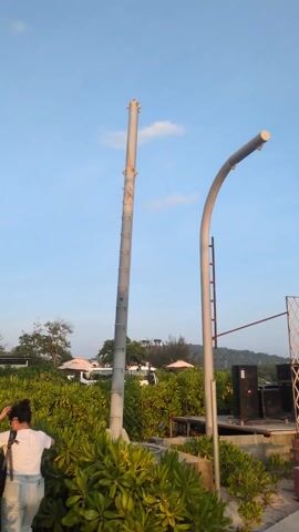 The video shows a beach scene with music playing. In the foreground are green bushes, two metal poles, one straight and one curved, and a small stage with speakers and a sign that says "DJ MUSIC BAR". In the background are trees, umbrellas, and a hill. The sky is blue with a few clouds.