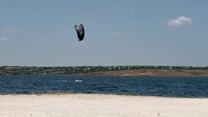 A kitesurfer is shown on a body of water. The kitesurfer is on a board and being pulled by a kite. The kite is dark colored with a logo on it. The water is blue and choppy. In the background, there is a shoreline with trees and buildings. The sky is blue with some white clouds. The foreground is a beach with white sand.