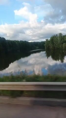The video begins by showing a calm lake bordered by a dense forest. The sky is partly cloudy with a blue sky visible through the clouds. The water reflects the sky and the surrounding trees. A metal guardrail is visible in the foreground, suggesting the view is from a moving vehicle. 

As the video progresses, a wooden structure with stairs is briefly visible in the lake, appearing to be a diving platform. The platform is partially submerged in the water. The vehicle continues to move, and the landscape shifts, showing more of the forested shoreline. The trees are a mix of deciduous and coniferous species, with lush green foliage. The water remains calm, with reflections of the sky and trees continuing to be visible. The video ends as it shows more of the forested shoreline passing by.