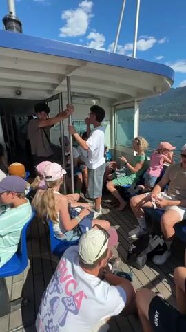 The video shows a group of people, a mix of adults and children, sitting and standing on the deck of a boat. The boat appears to be moving, as indicated by the water and distant shoreline visible through the windows. The deck is made of wood, and there are blue plastic seats arranged in rows. Several individuals are holding onto metal poles for stability. Two teenage boys are standing near the center of the frame, one holding onto a pole and the other looking towards him. In the foreground, a person wearing a white t-shirt with a mushroom graphic is visible from the back. Other passengers are seated, some looking at their phones or conversing. The overall atmosphere seems relaxed and casual, suggesting a recreational outing on the water under a bright, sunny sky with scattered clouds.