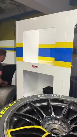 The video shows the inside of what appears to be a garage or workshop area associated with motorsports, possibly Formula racing. The scene begins focused on a used racing tire with "Pirelli" branding, positioned near a white cabinet with blue and yellow stripes. The camera pans to reveal a large, black-covered object, potentially a race car, and then moves past more cabinets and equipment. Finally, it reveals a control room-like space where two men are working at desks covered with multiple computer screens. The screens display technical data, and one of the men is adjusting his hair. They both wear black shirts with a logo featuring the same blue and yellow stripes. The walls of the room are yellow.