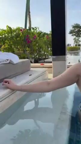The video shows a woman in a black and white striped bikini standing in a pool of clear water. She is leaning against the edge of the pool with her right hand. Her hair is pulled back in a bun. She is wearing a necklace with a key pendant and a black watch on her left wrist. The pool has a dark blue tiled bottom and a light grey tile trim. Behind the woman is a black pillar and a green bush with pink flowers. In the background, there are other trees and a blue sky with some clouds. The woman looks to her right, then looks at the camera and smiles.