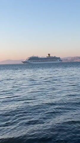 The video captures a serene seascape featuring a large white cruise ship with a yellow funnel, moving from right to left across the frame. In the distance, a shoreline with mountains is visible, softened by the atmospheric conditions. The sky displays a gentle gradient, transitioning from light blue at the top to a soft pale orange and pink near the horizon, indicative of either sunrise or sunset. The water is calm with gentle ripples. As the cruise ship exits the frame, a smaller boat appears further in the mid-ground, and a bird can be seen flying high in the clear sky. The overall scene conveys a peaceful moment on the sea, with the specified location context being Narlıdere, Turkey.