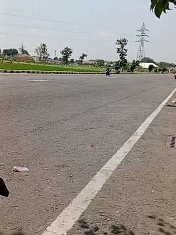 This is a landscape video taken from the side of a paved road. The road has two lanes in each direction and is bordered by a white line on the right side. There is a grassy median with some trees and bushes on the left side of the road. In the background, there are power lines and a tall transmission tower. A few motorcycles can be seen on the road, moving away from the camera. There is some litter on the side of the road, including a plastic bottle. The sky is clear and the sun is shining.