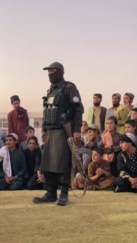 A man in tactical gear, wearing a mask and cap, stands holding a rifle in front of a crowd of people, many of whom appear to be children. The people are seated on the ground, some are clapping. The man in tactical gear appears to be addressing the crowd. The background suggests an outdoor event with a building in the distance. The lighting indicates it is likely late afternoon or early evening.