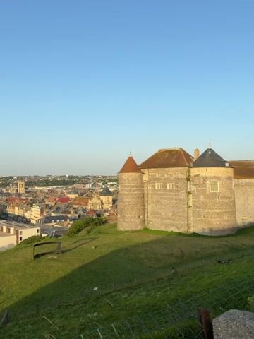 This video shows an aerial view of a coastal city, possibly taken from a hill overlooking the town. In the foreground, there is a grassy hill with a stone building that appears to be a castle or a historic structure. The building has circular towers and is made of stone. The city stretches out in the background, with buildings of various sizes and styles, some appearing to be residential and others commercial. A road runs through the city, with cars visible on it. The sea can be seen in the distance. The sky is clear and blue, indicating good weather. The lighting suggests it is either early morning or late afternoon, as there are long shadows cast by the objects on the hill.