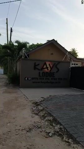 The video shows a dirt road leading away from the viewer. The road is flanked by walls and palm trees. On the right, a sign reads "KAYS LODGE" with contact information. The weather appears sunny and clear. The overall impression is of a quiet, tropical neighborhood.