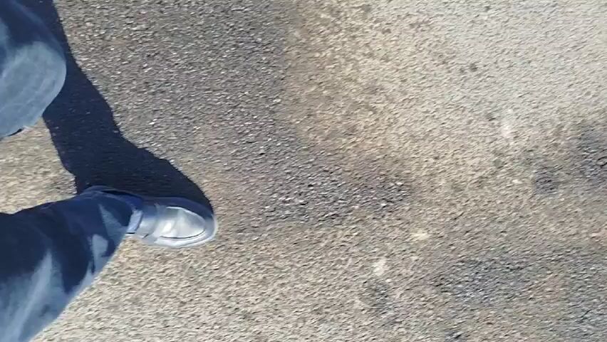 The video shows a person walking on asphalt pavement, casting a shadow. The video then shows a view of a street with multiple vehicles parked along the side, along with industrial buildings and power lines in the background. The sky is clear and blue.