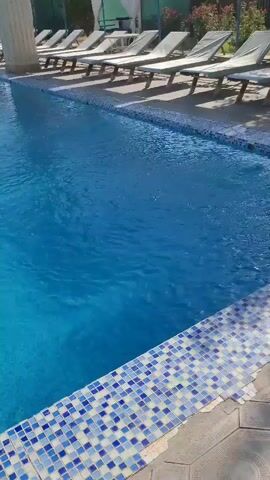 A blue swimming pool is shown, with lounge chairs lined up behind it. The pool has a tiled edge. The water is disturbed and appears to be moving. Sunlight is visible, suggesting it is daytime.