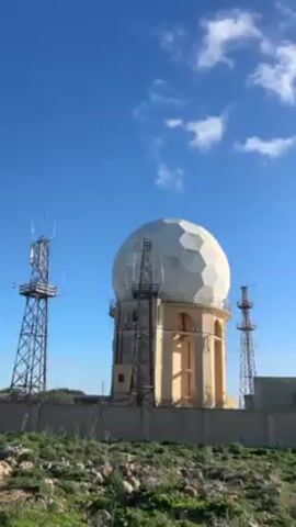 The video shows a white geodesic dome on top of a light yellow cylindrical tower, surrounded by tall metal lattice towers, behind a concrete wall. The sky is blue with some white clouds. The ground is covered with green vegetation and rocks. The camera pans to the right to show the sea in the background, with the sun shining brightly above it.