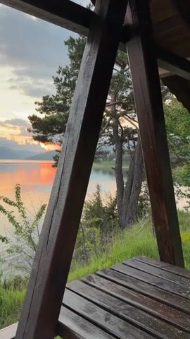 The video opens with a shot of a beautiful sunset over a lake, framed by wooden structures that appear to be part of a porch or balcony. The camera pans slowly to the left, revealing more of the stunning scenery. The sky is a mix of blues, grays, and oranges, with the sun's warm glow reflecting on the calm water. The surrounding landscape features lush green trees and distant mountains, creating a picturesque view. The video ends with a serene shot of the lake and the fading sunlight.