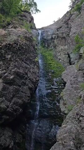 The video shows a narrow, vertical waterfall cascading down a rocky gorge. The rocks are rough and textured, with patches of green moss or vegetation growing on them. The water is clear and flows in a thin stream, breaking into smaller streams and droplets as it falls. The surrounding environment appears to be natural and wild, with trees visible at the top of the gorge. The lighting suggests it is daytime, possibly overcast or late in the day. There are no people or man-made objects visible in the frame.