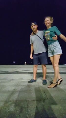 A man and a woman are standing on a dark, paved plaza. The man is wearing a gray t-shirt, navy blue shorts, a dark baseball cap, and navy blue Crocs. He has a gray satchel slung over his left shoulder. The woman is wearing a teal t-shirt, jean shorts, and black sandals. The man lifts the woman into his arms and spins her around. The background is black, with some lights visible in the distance.