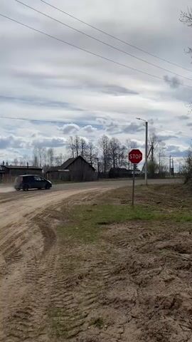 The video shows a car speeding down a dirt road. The car, which is dark gray or black, kicks up a cloud of dust as it drives. There is a stop sign at the intersection. The environment is rural, with a few houses visible in the background, and trees without leaves. The sky is overcast.