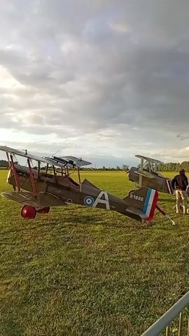 Two vintage biplanes, one a brown military aircraft with a white "A" and a blue and white circular insignia, the other with French national markings on its tail, are parked on a grassy field under a cloudy sky. A person in dark clothing stands near the tail of the second biplane. The scene appears to be an outdoor event or display.
