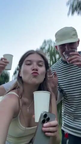 Here is a description of the video.
The video shows three young adults toasting. The woman in the foreground holds a white paper cup and an iPhone. She has long brown hair and wears a white top. To her right is a man in a black and white striped shirt and a tan cap. He is holding a cigarette and a clear plastic cup with berries. Behind the woman is another young woman in a white shirt holding a paper cup high in the air. They appear to be at an outdoor gathering. Trees and grass are visible in the background. The sky is light.
