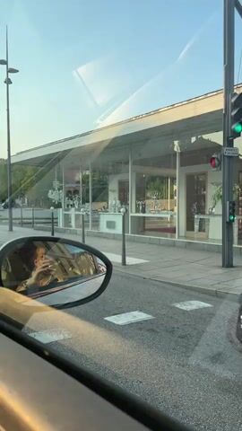 A person's reflection is seen in a car's side mirror, holding a drink. The car is driving past a building with large glass windows displaying chandeliers and other decorative items. The building appears to be a shop. Traffic lights are visible, with one showing a green light. The scene transitions to show the interior of the car, with the dashboard and steering wheel visible. The car is driving down a street lined with buildings, and a roundabout is visible in the distance.