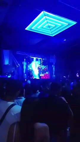 The video shows a crowded indoor space with a stage at the far end, brightly lit with blue and white lights. A man in a suit stands on the stage, appearing to speak or sing into a microphone. The stage has a large screen behind him, displaying a colorful, abstract design. Two statue-like figures stand on either side of the screen. The ceiling features a square, tiered light fixture that emits a blue glow. The audience consists of numerous individuals, most of whom have their backs to the camera. Some audience members are wearing white shirts. Overall, the atmosphere suggests a concert or performance event.