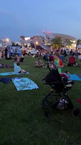 The video shows a crowd of people gathered on a grassy area in the evening. They are seated on blankets and lawn chairs, with some standing. In the background, a stadium structure with lights is visible, along with trees and tents. A stroller is in the foreground. The sky is a dusky blue. The people are casually dressed and appear to be socializing or waiting for an event. The overall atmosphere is relaxed and communal.