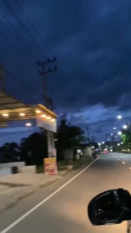 The video shows a road at dusk or night, from the passenger perspective of a moving vehicle. The sky is a deep blue, filled with dark, heavy clouds. Streetlights illuminate the road ahead, and there are utility poles and wires along the roadside. Small buildings and shops line the side of the road, and some vehicles are visible in the distance. The side mirror of the vehicle is visible in the foreground, showing the reflection of the passing scenery. The wind is causing significant noise.