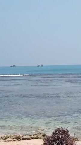 The video shows a serene ocean view with three boats visible on the horizon. The water is calm, with gentle waves lapping at the shore. The sky is clear and blue, indicating a sunny day. The foreground features a sandy beach with some rocks and debris. The overall atmosphere is peaceful and tranquil.
