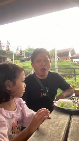The video shows a man and a little girl sitting at a wooden picnic table under a covered area. The man is wearing a black t-shirt with a palm tree design on it and has a strap slung across his chest. He has short, dark hair and is looking directly at the camera. The little girl, who is sitting to his left, is wearing a pink ruffled top and has long, dark hair. She is eating something and looking at the camera. On the table in front of them is a plate of food, which includes rice, skewers, and lettuce. The background of the video shows a grassy hill with a building on top. Other people are walking around in the background. The lighting is bright, suggesting it is daytime.