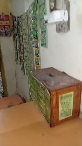 A video shows a wooden donation box with text on it in a room. The donation box is placed on a table or counter. To the left of the donation box, there are rows of small packets hanging on the wall. The packets appear to be food items, possibly snacks or spices, and they are predominantly green in color. To the right of the donation box, a white electrical fixture with multiple outlets is mounted on the wall. The wall itself is painted in a light color and shows some signs of wear and discoloration. The flooring visible in the background is tiled with a geometric pattern. The lighting in the room is dim.