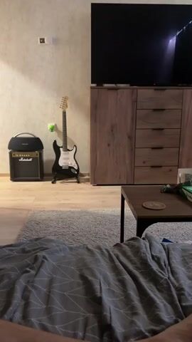 The video shows a room with a television on a wooden cabinet. To the left of the cabinet, a black electric guitar stands on a stand. Next to the guitar is a Marshall amplifier. In the foreground, a person is lying under a gray blanket. The camera pans slightly to the right, revealing a coffee table with a board game on it. A teal curtain is visible in the background.