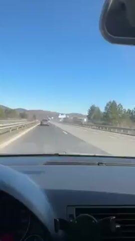The video shows a driver's perspective while driving on a highway under a clear blue sky. There are other cars on the road, and trees and hills can be seen in the distance. The dashboard of the car is visible in the foreground.