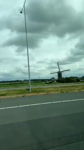 The video shows a Dutch windmill with its blades turning, seen from a moving vehicle traveling on a road. The windmill is green with a brown base, and its blades are a dark color, possibly black or dark brown. It is surrounded by lush green grass and trees. In the background, there are more trees, some buildings, and a cloudy sky. The road in the foreground has white lines marking the lanes. The video is shot from a slight angle, and the movement of the vehicle causes a slight blurring of the scenery.