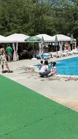 A poolside scene is depicted. The camera is positioned at ground level, looking towards a swimming pool. The pool is filled with bright blue water and has lounge chairs arranged around it. Several white canopies with green accents provide shade. Trees line the background. In the foreground, a woman in a white and black outfit is sitting on the concrete with two young girls. One girl, wearing a white and pink outfit, is also sitting. Another girl in a pink swimsuit is kneeling at the edge of the pool. A man in a gray t-shirt and shorts stands near the girl in the pink swimsuit, facing the pool. Other people are visible in the pool and further back, lounging or walking. The ground in the immediate foreground is covered with green artificial turf.
