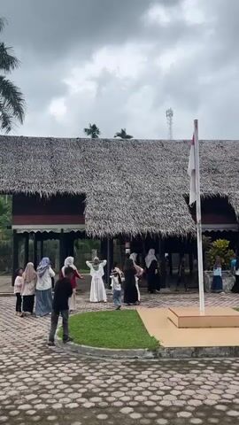 In an outdoor setting with a cloudy sky, a group of people are gathered in front of a building with a thatched roof. Some people are walking, while others appear to be observing or interacting. There is a flagpole with a flag flying. The ground is paved with decorative stones. Palm trees are visible in the background, suggesting a tropical or subtropical environment. The people are dressed in casual clothing, and some are wearing hijabs. The scene appears to be a public gathering or event.