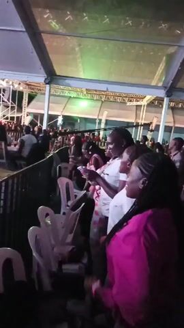 The video shows a crowd of people at an event under a large tent structure. A group of women are standing close to the camera, clapping and enjoying the music. There are numerous white plastic chairs stacked nearby. Other people in the background are standing, talking, and some are holding phones. The tent's ceiling is visible, supported by a metal frame and lit by colorful stage lights. The overall atmosphere appears lively and festive.