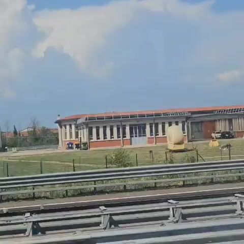 The video shows a highway scene from the perspective of a moving vehicle. In the foreground, metal guardrails are visible. Beyond the guardrails, various trucks are seen driving on the highway. One truck is white with a blue trailer. Another truck has a grey trailer. A third truck is white with a blue and red logo. In the background, there is a brick building with a row of windows and a red roof, situated behind a grassy field. A large, light-colored, abstract sculpture is in the field. Trees and bushes are also visible in the background. The sky is blue with scattered clouds.