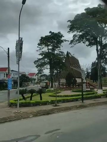 The video captures a view of a park area from what appears to be a moving vehicle. The park features a distinctive building with a thatched roof, surrounded by manicured greenery, trees, and decorative white structures resembling trees with illuminated elements. In the background, there is a monument or statue. The sky is overcast with clouds, suggesting a possibly cool or damp day. The road in the foreground shows signs of wear, with a curb and drainage elements. There are trash receptacles visible near the entrance of the park. The overall impression is a public space with unique architectural features and landscaping.