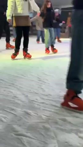 The video shows several people ice skating on an ice rink. The camera focuses on the lower body of a person wearing a white jacket, brown skirt, and black leggings, along with a child wearing a black jacket and jeans, both wearing orange skates. They are moving around the rink with other skaters, illuminated by multicolored lights. The ice is marked with skate tracks.