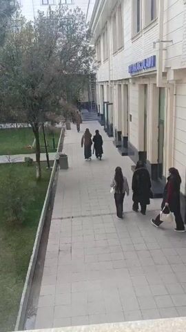 The video shows a sidewalk scene with a building on the right side and a grassy area with trees on the left. Several people are walking along the sidewalk in the same direction. The building has a sign written in a language other than English. Some of the people are wearing dark clothing. There are lampposts along the sidewalk. The sidewalk is made of light-colored square tiles. A ladder is visible along the building.