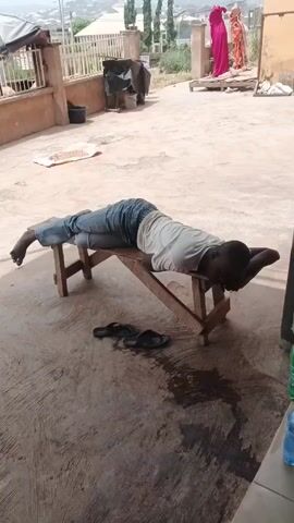The video shows a person lying prone on a small wooden bench outdoors. The person is wearing a light-colored shirt and jeans. Their hands are behind their head. A pair of sandals is on the ground beside the bench. There are two people standing in the background near a white railing; one is wearing a pink garment and the other a patterned one. The ground is concrete and has a few dark stains. Behind the railing are several buildings and trees. There is a white sheet or cloth on the ground to the left of the bench.