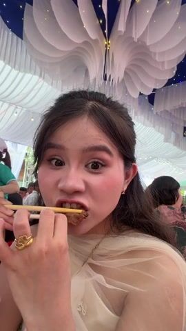 A young Asian woman with dark hair is eating something with chopsticks in a selfie-style video. She is wearing a light-colored dress and gold jewelry. She looks directly at the camera as she bites into the food, then chews with her mouth closed. Other people are visible in the background, some in suits, suggesting a formal event. The background also shows a white tent with decorative white fabric hanging from the ceiling.