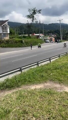 The video shows a cyclist riding on a highway. The cyclist is wearing a white and red suit, with a helmet. The cyclist is riding to the right in the frame, on the side of the highway that is closest to the camera. There is a median on the highway separating the direction of traffic.  In the background, to the left, is a small white house and a tree. In the distance, mountains can be seen. To the right of the cyclist, there is a person standing off the side of the highway, as well as a police officer on a motorcycle. There are a couple other people standing in the distance as well. As the cyclist continues down the highway, a dark blue car passes in the opposite direction. There is a large billboard on the side of the highway.  The weather appears to be overcast.