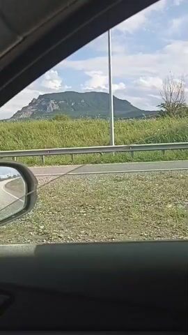 The video shows a view from inside a vehicle looking out the window. The scene outside is a rural landscape with a road, a guardrail, and a grassy field. In the distance, there are hills and mountains under a cloudy sky. The camera is moving, indicating the vehicle is in motion. At one point, a road sign is visible on the left side of the road. The video is short and appears to capture a moment of travel through a natural setting.